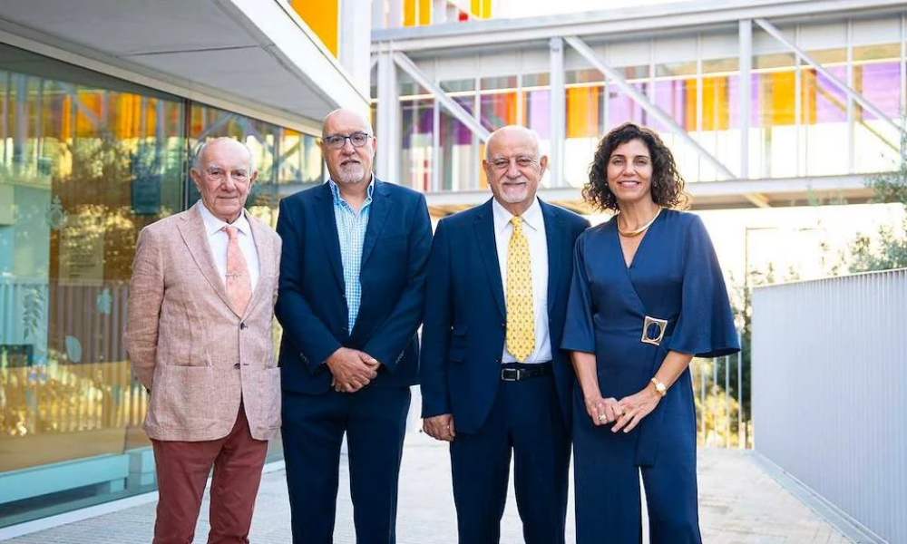 Fotografía de grupo tomada en las instalaciones del Hospital Sant Joan de Déu Barcelona. Cuatro profesionales posan de pie frente a un edificio moderno con pasarelas acristaladas y elementos arquitectónicos en tonos amarillos y blancos, en el marco del Simposio Internacional sobre Cardiopatías Hereditarias Pediátricas.