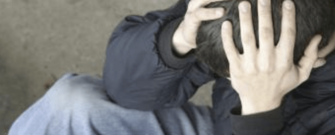 Imagen de un adolescente, sentado en el suelo, sujetándose la cabeza. Aparentemente parece preocupado, mirando hacia el suelo.